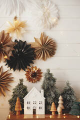 Stylish christmas little house, trees, golden lights on background of paper stars on white wooden...