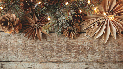 Stylish christmas paper stars, fir branches, cones and lights border on rustic old wooden background. Top view with space for text. Season's greeting card. Merry Christmas and Happy Holidays!