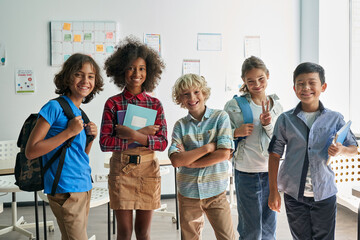 Portrait of cheerful smiling diverse schoolchildren standing posing in classroom holding notebooks and backpacks looking at camera happy after school reopen. Diversity. Back to school concept. - Powered by Adobe