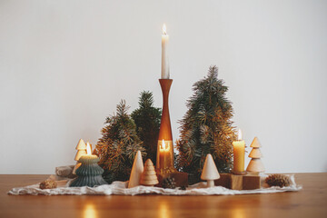 Stylish christmas candles and pine trees decorations on wooden table on background of white wall in...