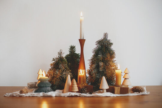Stylish christmas candles and pine trees decorations on wooden table on background of white wall in modern scandinavian festive room. Atmospheric winter time. Holiday advent