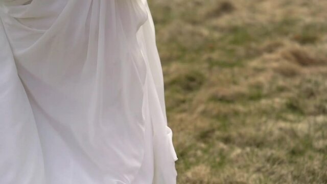 The blonde bride walks across the field, picks up and holds her long white dress. Wedding on a sunny day in the countryside.