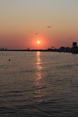 Fototapeta premium Sunset paints the sky and sea in shades of red. Seagulls fly against the backdrop of the setting sky and the setting sun. The sea pier is visible in the distance. The resting people watch the sunset