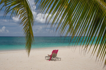 A sole chaise lounge chair sits on an empty beautiful clear and turquoise water beach on the Cayman Islands near a small grove of palm trees