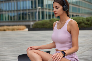 Photo of relaxed woman in sportsclothes sits crossed legs on fitness mat listens relaxing music practices yoga in downtown enjoys relaxation and meditation. Urban background. Healthy lifestyle