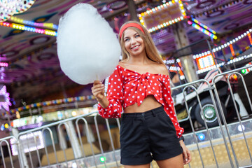 Adorable cute beautiful woman with cotton candy stands in the middle of an amusement park with bright colors, positive and cheerful, happy and optimistic emotion