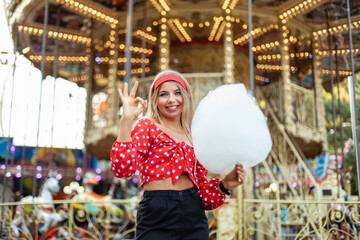 Obraz na płótnie Canvas Adorable cute beautiful woman with cotton candy stands in the middle of an amusement park with bright colors, positive and cheerful, happy and optimistic emotion