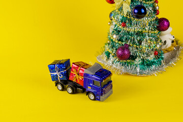 Toy truck carrying boxes of Christmas presents. Blue car with colorful boxes. Christmas tree on yellow background.