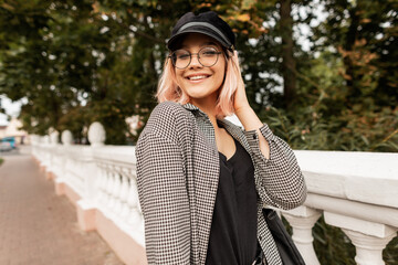 Happy beautiful young teenager girl with cheerful emotions and a smile in fashionable casual clothes with glasses and a cap walks on the street