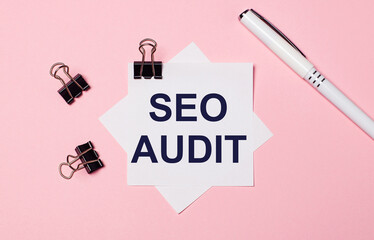 On a light pink background, black paper clips, a white pen and white note paper with the text SEO...