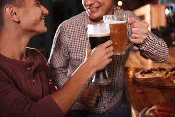 Cropped shot of cheerful male friends clinking beer glasses, celebrating at the bar