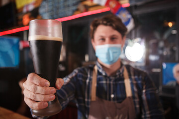 Close up of a professional brewer wearing medical face mask, holding beer glass to the camera
