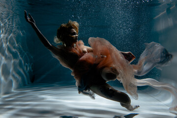 A girl with long dark hair swims underwater in a pink dress and with a crown on her head, like an underwater queen. Fairy tale suitable for advertising	