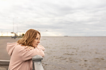 Fototapeta na wymiar A young woman on a cloudy evening stands on the embankment and looks at the sea