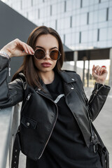 Obraz na płótnie Canvas Urban beautiful young cool girl with the sexy beauty face in fashionable round sunglasses with a stylish black leather jacket and dress with handbag walks on the street