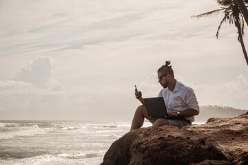 Cyfrowy nomada, człowiek pracujący z laptopem i smartfonem zdalnie na tle oceanu i skał...