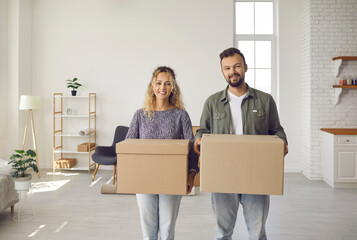 Portrait of happy young married couple who bought house standing in new home holding boxes, looking at camera. Two people support modern charity project, give things, donate stuff, help social service