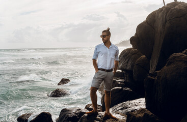 Mężczyzna elegancko ubrany stojący na skałach na tle oceanu i wzburzonych fal.