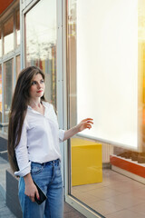 Fototapeta na wymiar Young happy woman standing near luminous shop window and looks at place of mockup advertising
