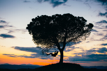 Fototapeta na wymiar Silhouette of a young woman enjoying freedom at sunset. Tire swinging from a tree outdoors. Holiday concept.