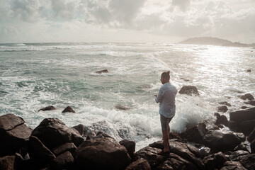Mężczyzna elegancko ubrany stojący na skałach na tle oceanu i wzburzonych fal. - obrazy, fototapety, plakaty