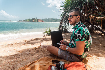 Cyfrowy nomada, mężczyzna pracujący na tropikalnej plaży z laptopem i telefonem. - obrazy, fototapety, plakaty