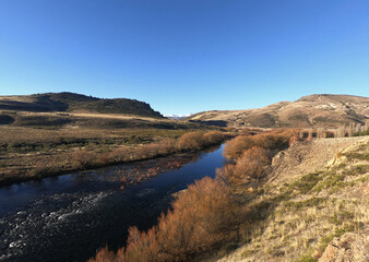 Idyllic rural landscape with a clear blue sky. The river flowing across the golden valley, yellow grassland, forest and mountains in autumn. 