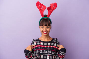Young mixed race woman with reindeer hat celebrating Christmas isolated on purple background  person pointing by hand to a shirt copy space, proud and confident © Asier