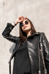 Young beautiful female model girl in fashionable sunglasses with stylish black leather jacket stands near the concrete gray texture wall on a sunny day in sunlight