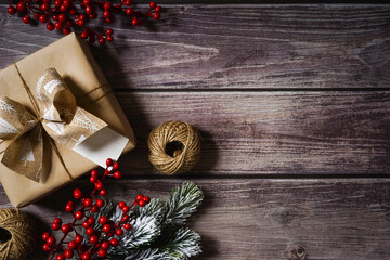 Christmas present wrapped in natural brown paper with decoration on dark wooden background. Festive holiday composition. Merry Christmas. Red berry, branches, twine. Empty space for text. Copy space.