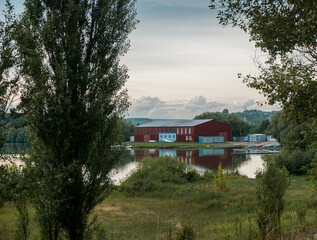 a red warehouse stands on the shore of a lake, trees are on the edges of the frame, a summer day