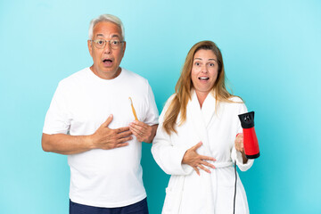 Middle age couple holding dryer and toothbrush isolated on blue background with surprise and shocked facial expression