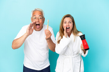 Middle age couple holding dryer and toothbrush isolated on blue background shouting and announcing something