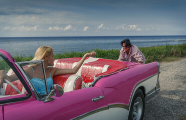 Young man pushing pink vintage convertible at coast whilst girlfriend drives, Havana, Cuba - obrazy, fototapety, plakaty