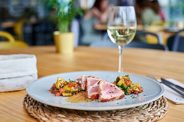 diner view of pork tenderloin dish with avocado salad accompanied by a glass of white wine at a restaurant table with people in the background out of focus.