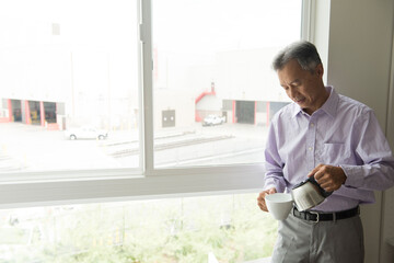 Mature man standing in front of window pouring hot water into teacup