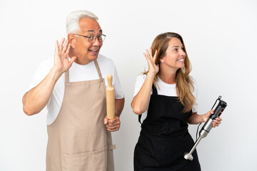 Middle age chefs couple isolated on white background listening to something by putting hand on the ear