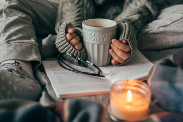 Female hands holding cozy mug with cacao or chocolate. Woman in gray knitted sweater sitting on bed. - 456580373