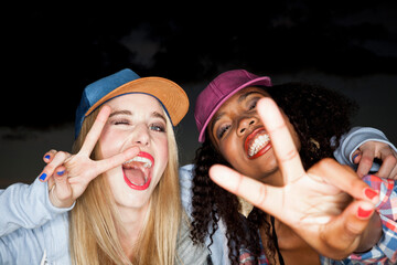 Young woman wearing baseball cap arm around friend looking at camera doing peace sign