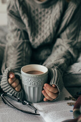 Female hands holding cozy mug with cacao or chocolate. Woman in gray knitted sweater sitting on bed.
