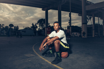 Man on basketball court sitting on basketball looking at camera