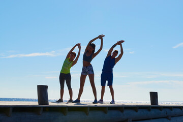 Friends on pier wearing exercise clothes arms raised, bending over sideways stretching