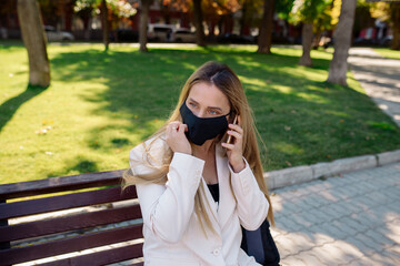 Young woman wearing a medical protective mask sits on a park bench during the coronavirus pandemic and talks on a mobile video