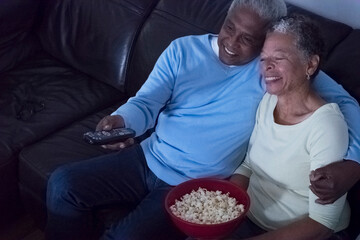 Senior couple sitting on sofa at night, watching television, woman holding bowl of popcorn