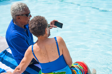 Senior couple sitting beside swimming pool, using smartphone