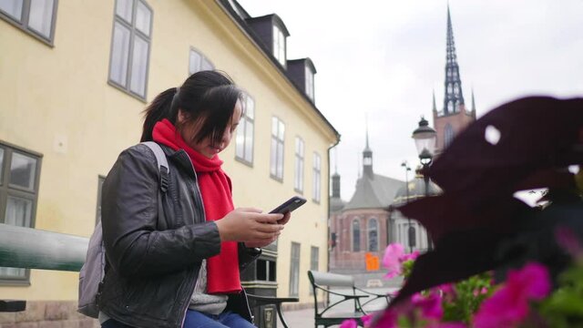 Asian woman wearing winter outfits sitting and using smartphone in town , going out for a walk to visit the city in winter on the street in Sweden. Traveling abroad on long holiday