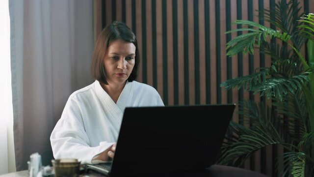 A girl in a white housecoat is sitting at a table and working at her laptop. A freelancer at work. The brunette performs the task of her clients. She took a job at home.