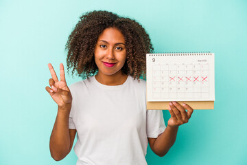 Young african american woman holding a calendar isolated on blue background showing number two with...