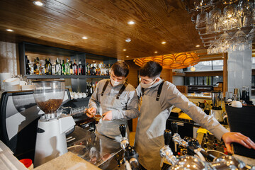 Two masked baristas prepare delicious coffee in the cafe bar. The work of restaurants and cafes during the pandemic.