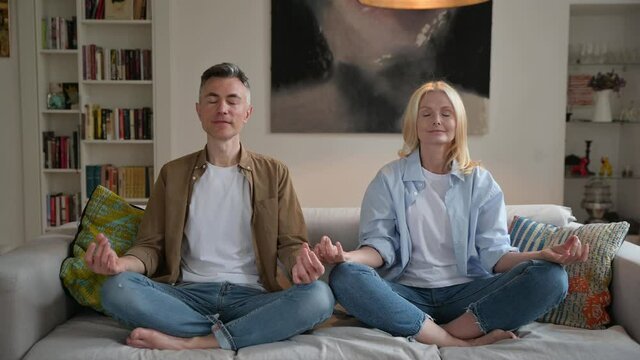 Middle aged married couple in casual clothes, leading a healthy lifestyle, doing yoga at home on the couch, sitting in lotus position in the living room, meditating, eyes closed, smiling. Calmness 